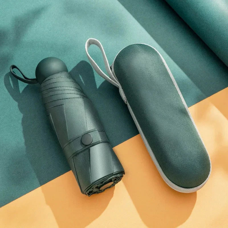 A compact, dark teal travel umbrella with a light gray fabric sleeve is shown on an orange surface.