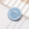 A round, light blue handmade PU leather label with a stitched heart design placed on a piece of parchment paper.