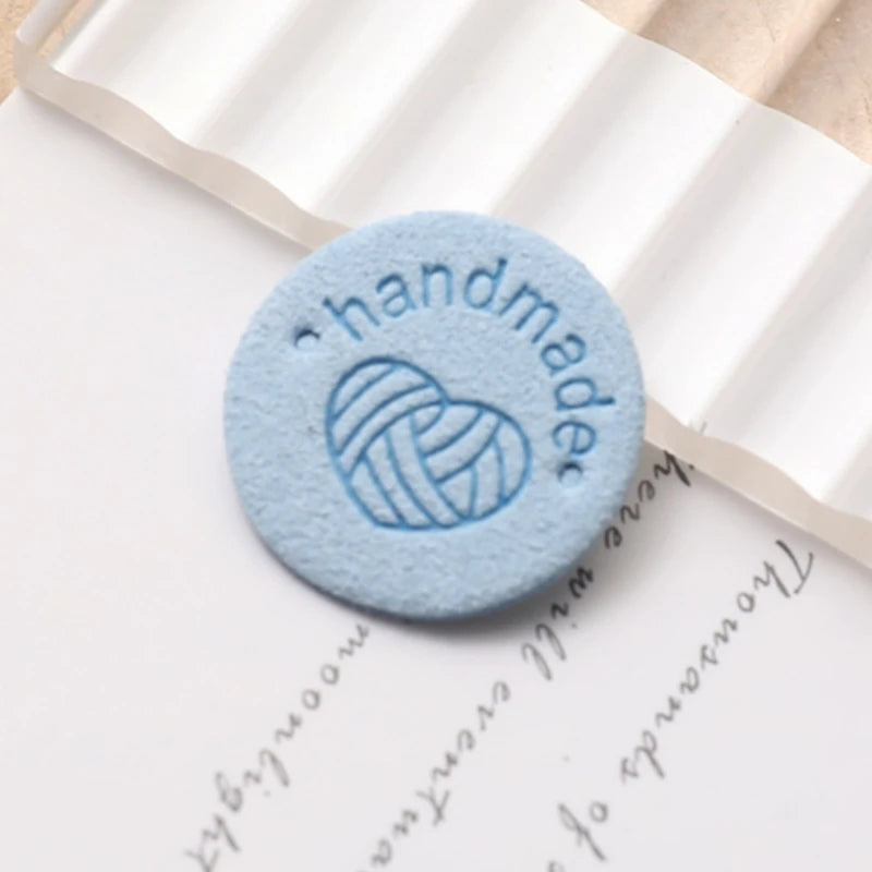 A round, light blue handmade PU leather label with a stitched heart design placed on a piece of parchment paper.