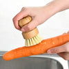 A hand is using a bamboo dish scrub brush with natural sisal bristles to clean an orange carrot in a metal bowl.