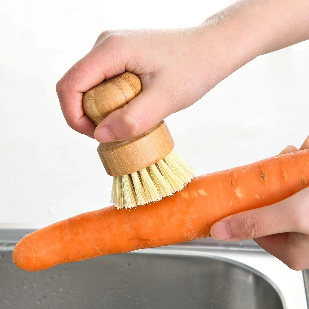 A hand is using a bamboo dish scrub brush with natural sisal bristles to clean an orange carrot in a metal bowl.