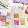 A clear acrylic quilting ruler with a dual scale grid and seam guide is shown alongside fabric squares and a sewing thread.