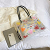A clear transparent tote bag with a black strap and an inner pouch filled with heart-shaped confetti.