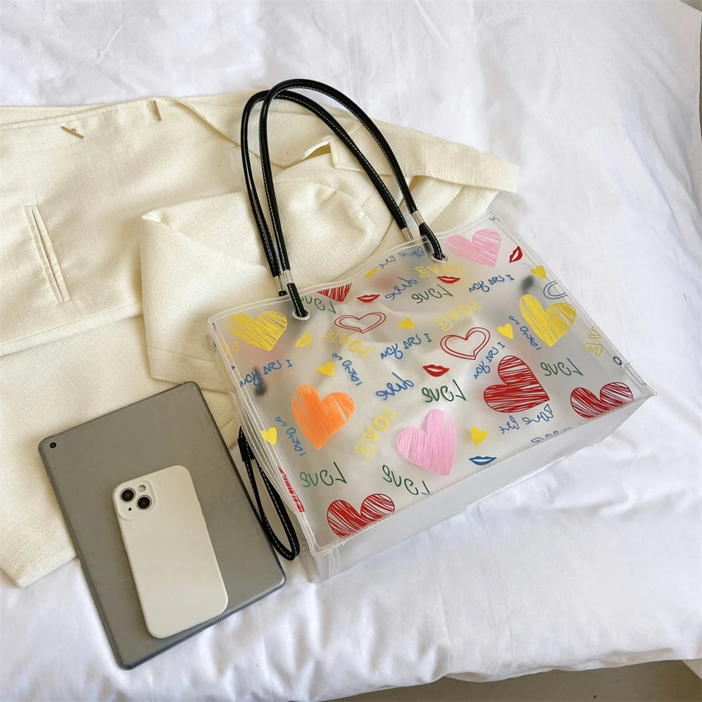 A clear transparent tote bag with a black strap and an inner pouch filled with heart-shaped confetti.