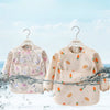 A light beige children's waterproof long sleeve feeding bib with a catch pocket displayed on a hanger against a blue sky back