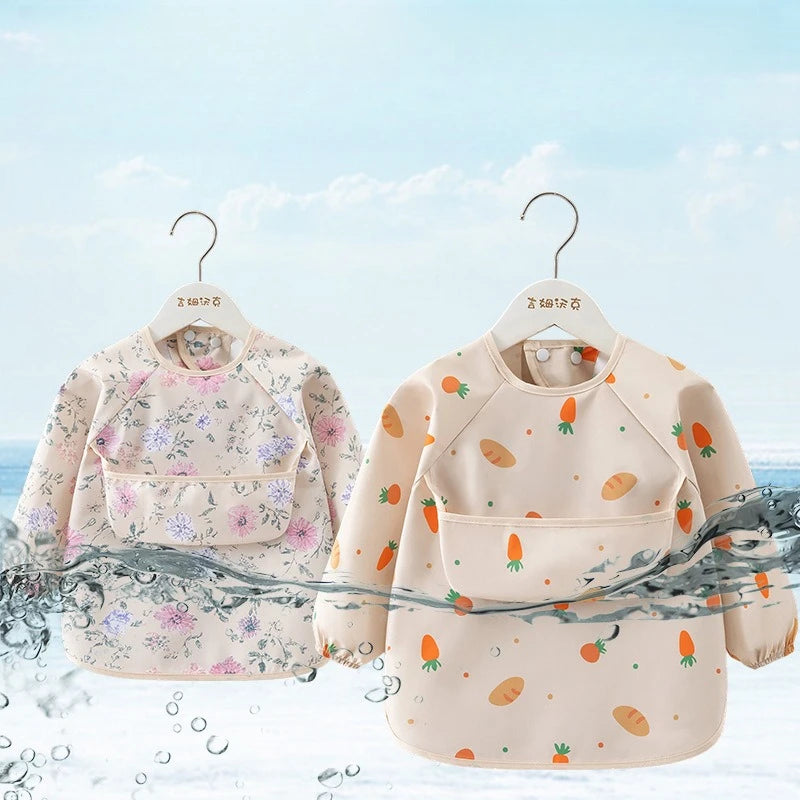 A light beige children's waterproof long sleeve feeding bib with a catch pocket displayed on a hanger against a blue sky back