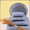 A stack of blue gray needle felting mats rests on a surface next to a bundle of golden wheat.
