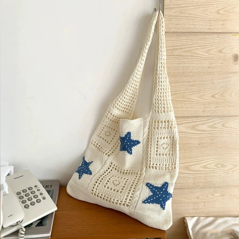 A cream-colored handwoven knit tote bag with a blue star pattern hanging next to a wooden surface.