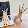 A beige resin hand planter and pen holder with wooden sticks is displayed next to a laptop.