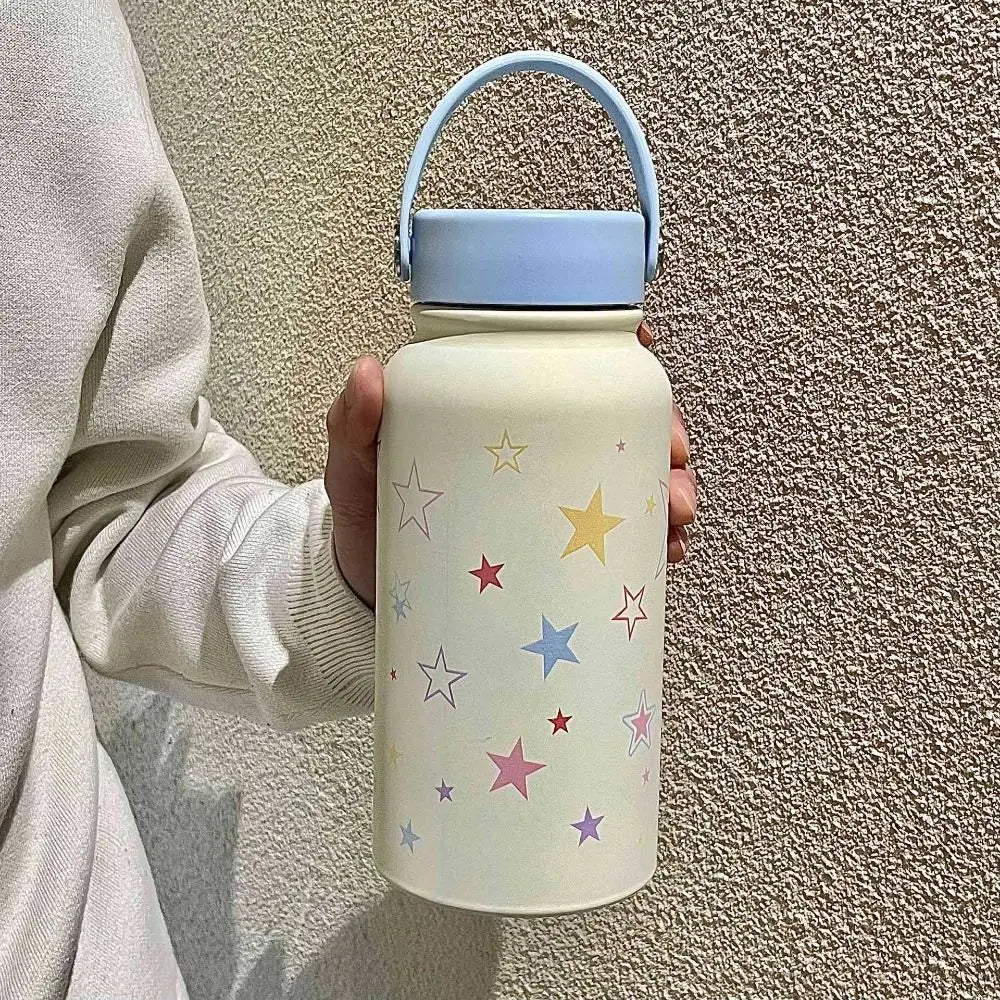 A hand holds a light beige stainless steel insulated travel tumbler with a blue lid against a textured wall.