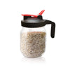 A clear glass jar filled with dried herbs or spices, topped with a black lid featuring a red pour spout and handle.