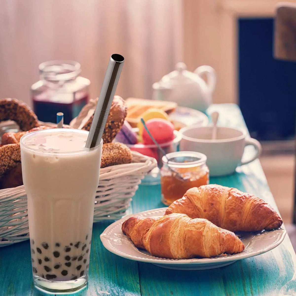 A glass of milk or smoothie is shown next to a reusable stainless steel drinking straw and various baked goods on a wooden ta