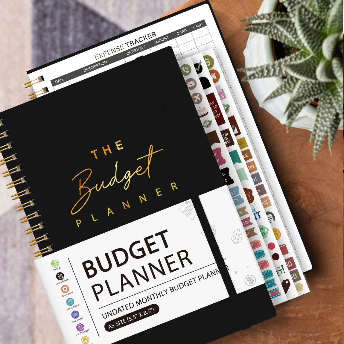 A spiral-bound budget planner with monthly and weekly pages and stickers is shown on a wooden surface.