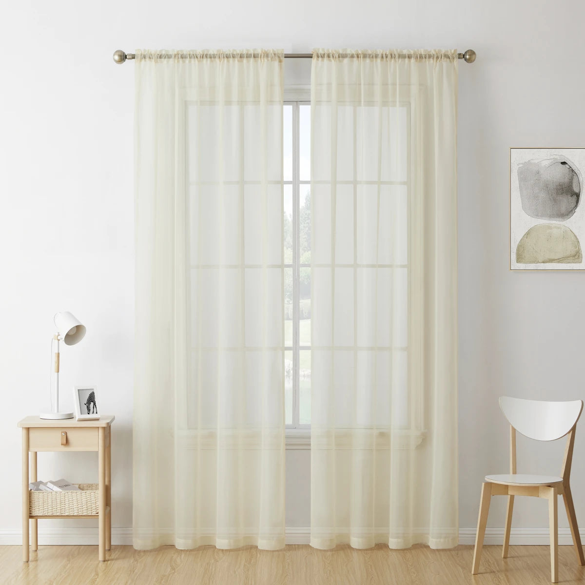 Two panels of sheer white voile curtains hanging in a window with a light wood table and chair nearby.