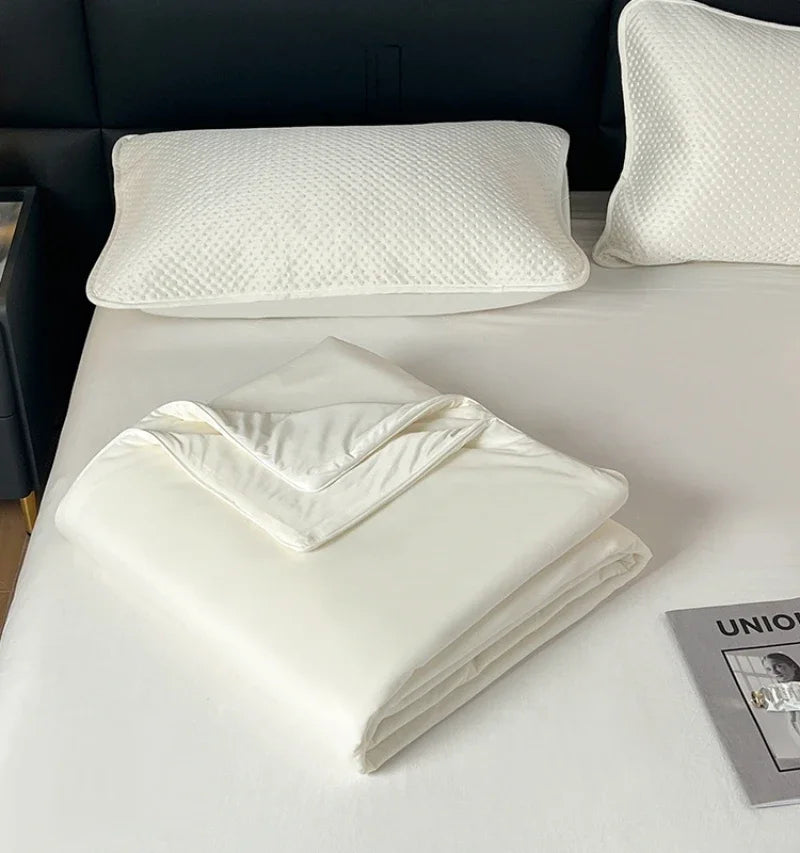 A white lightweight ice silk quilt is folded on a white bed with a white quilted pillow nearby.