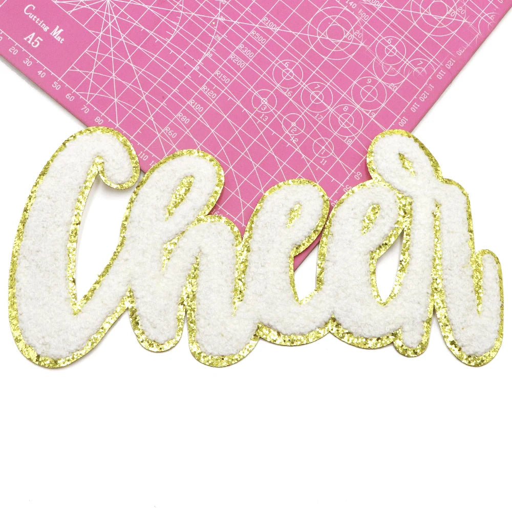 A gold glitter chenille letter patch spelling 'Cheer' on a pink cutting mat.