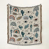 A woven throw blanket featuring a bohemian pattern of cacti, eyes, and rainbows on a light fabric with fringed edges.