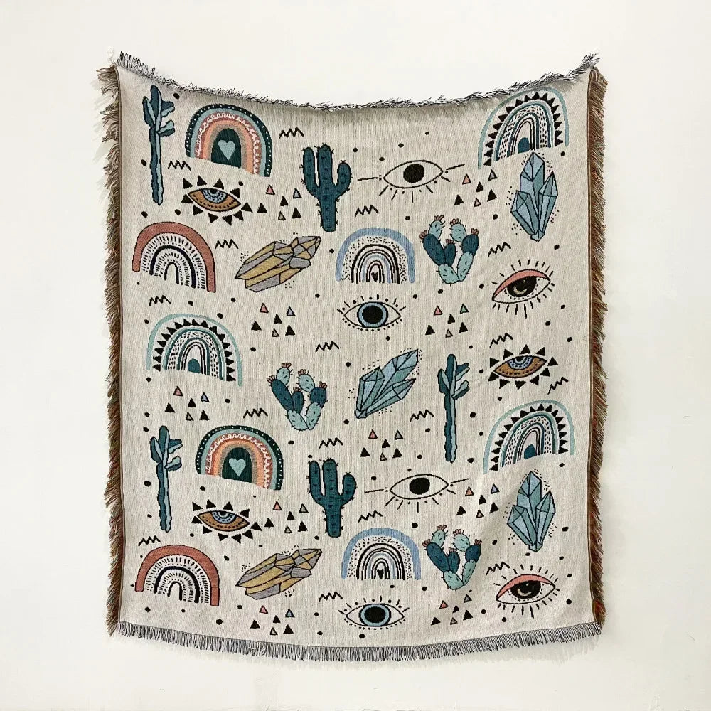 A woven throw blanket featuring a bohemian pattern of cacti, eyes, and rainbows on a light fabric with fringed edges.