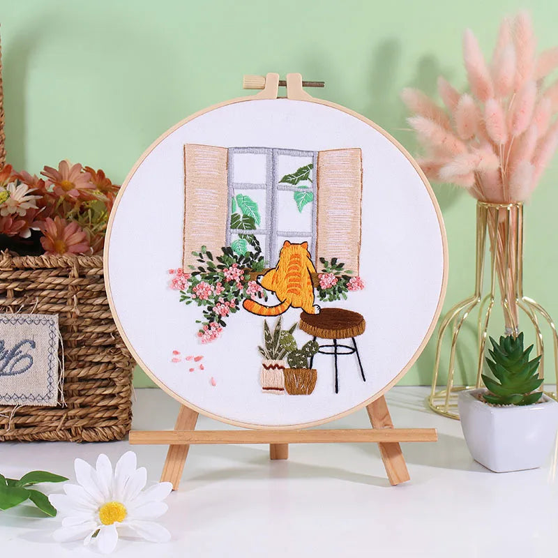 A circular embroidery hoop displaying floral and cat designs, set against a light green background.