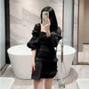 A woman in a black ruffled dress holds an elegant patent leather clutch bag with a chain strap.