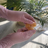 Eine Hand mit rosa Einweghandschuh hält eine orange Frucht in hellem Sonnenlicht.