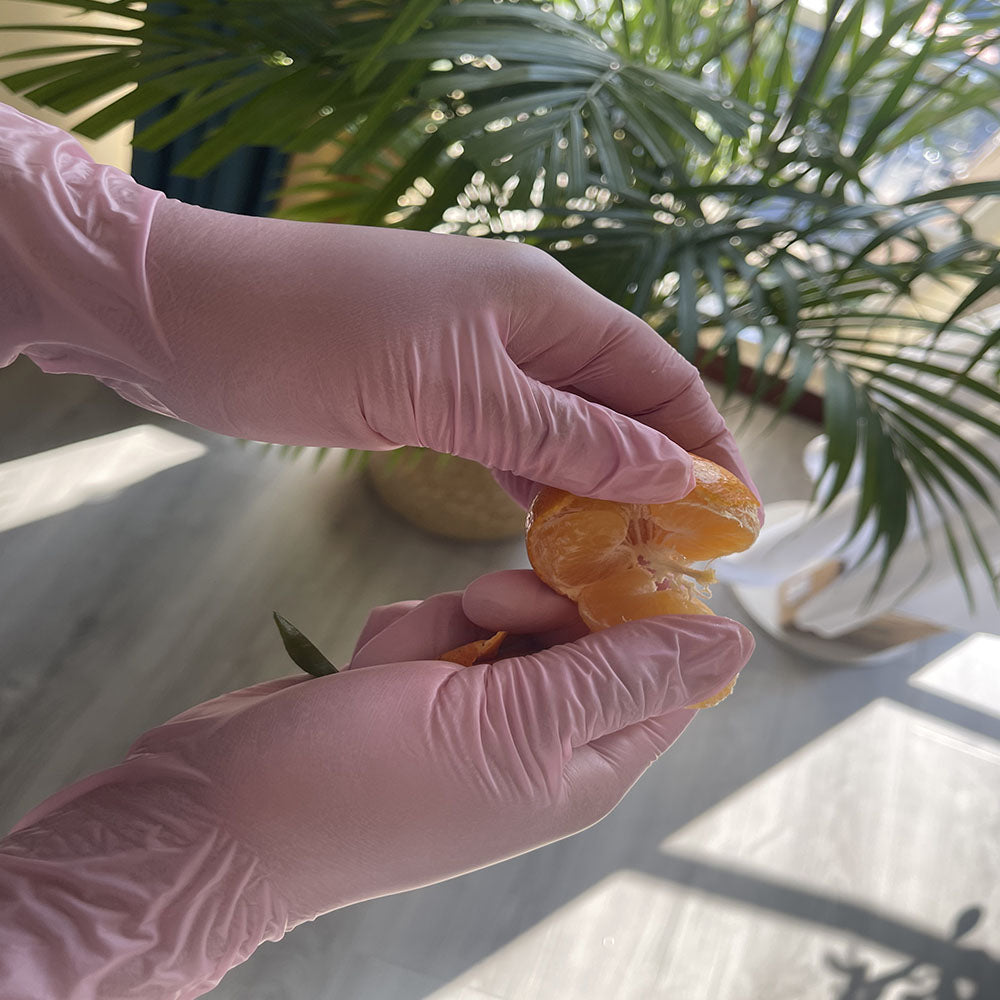 Eine Hand mit rosa Einweghandschuh hält eine orange Frucht in hellem Sonnenlicht.