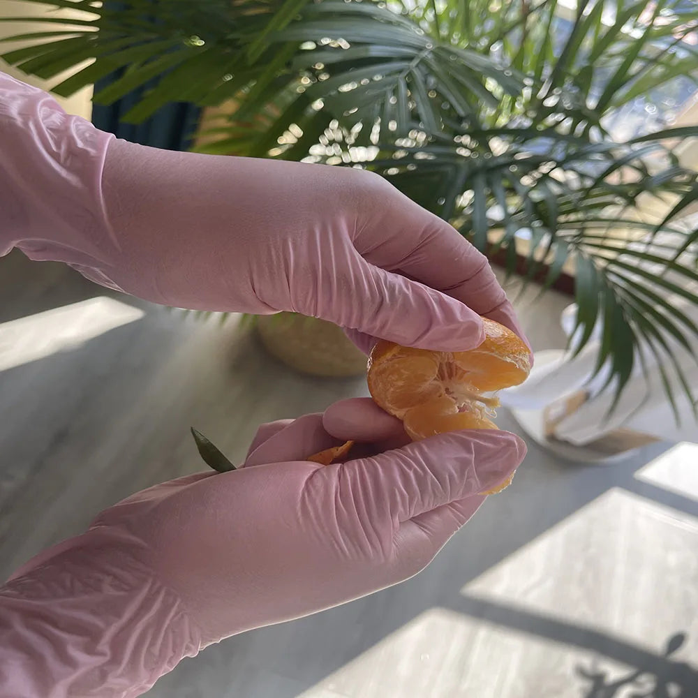 Eine Hand trägt eine pinke Einweghandschuh und hält eine orange Frucht bei hellem, natürlichem Licht.