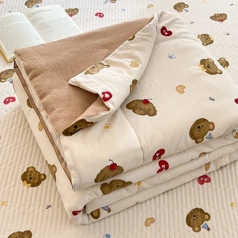 A beige quilt with a light pattern of cute brown bear and red heart motifs is folded on a white surface.
