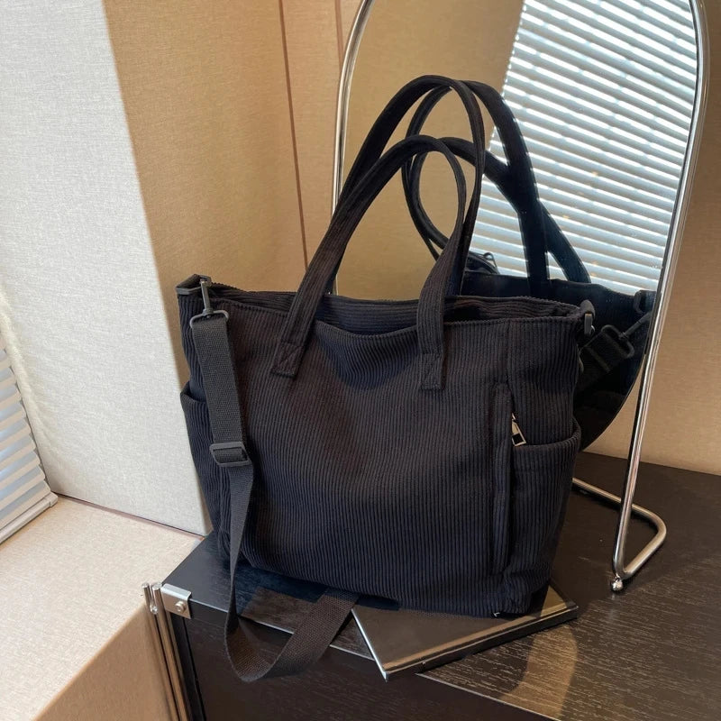 A black corduroy tote bag with an adjustable strap is displayed in front of a silver-framed mirror.