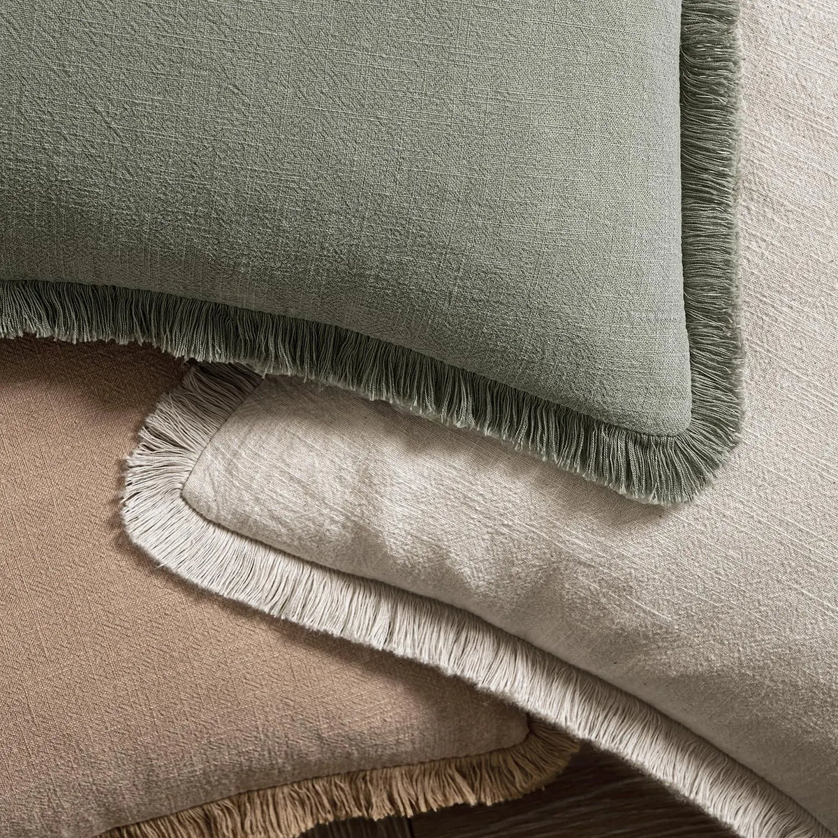A close-up of a sage green boho fringe throw pillow cover with a natural linen texture resting on a beige cushion.