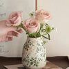Eine Hand hält eine Vase mit rosa Rosen in einer handgefertigten Eis-gecrackten Keramikvase mit grünem Laub.