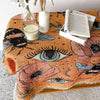 A bohemian jacquard woven throw blanket in orange and brown tones with embroidered eyes and floral patterns, displayed with c