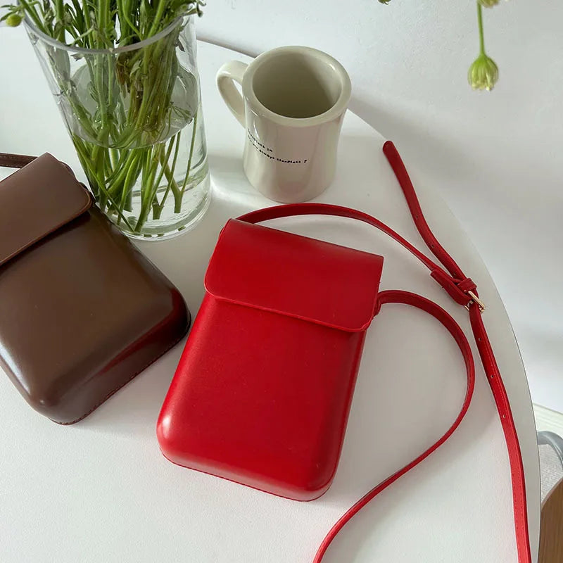 A bright red vintage PU leather crossbody shoulder bag resting on a white surface next to a glass vase and a ceramic mug.