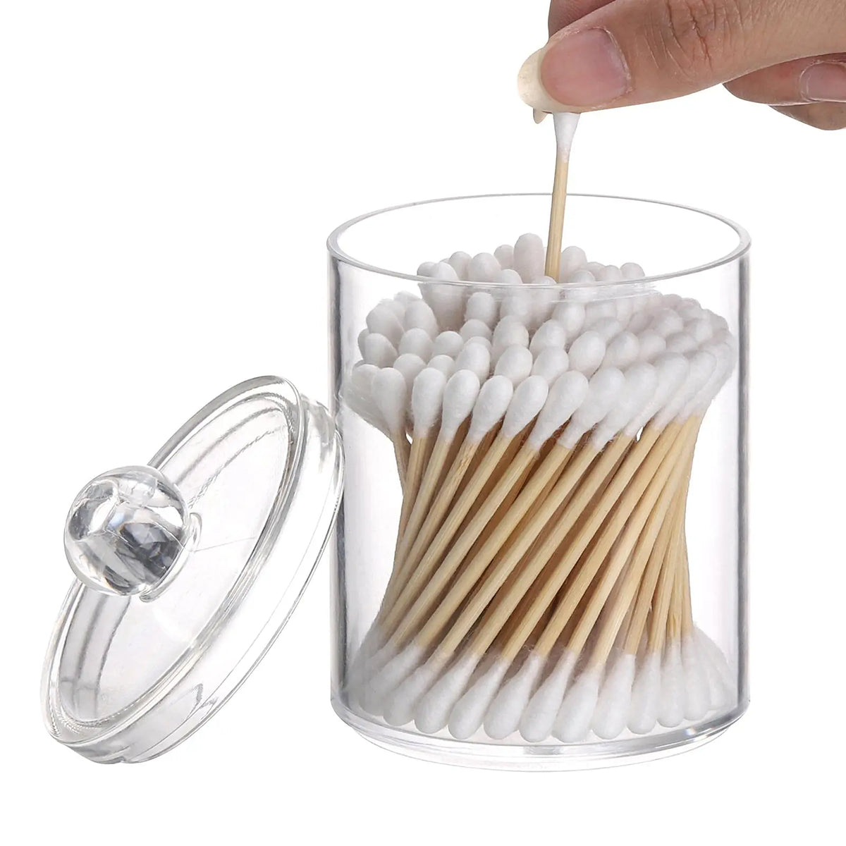 A hand dips a white cotton swab into a clear glass jar filled with numerous swabs and wooden sticks.
