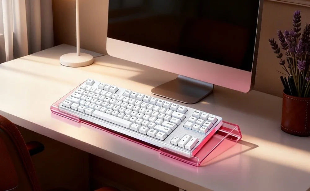 A pink acrylic ergonomic keyboard riser with a non-slip base is placed on a light desk next to a computer monitor.