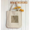 A beige canvas tote bag with a geometric stencil design and a bunch of yellow flowers hanging on a wooden rack.