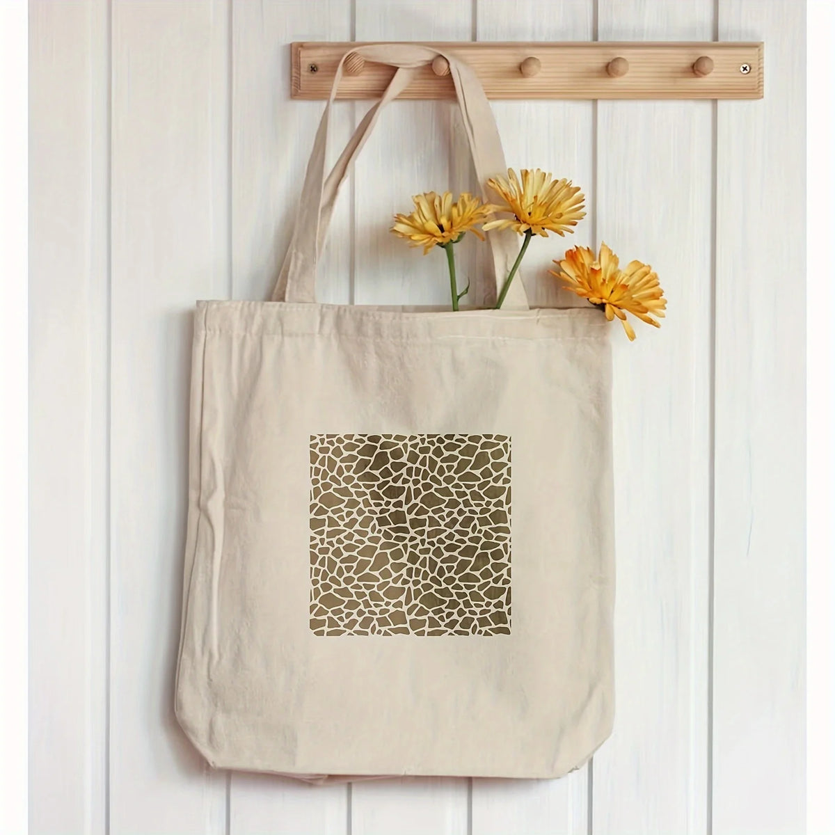 A beige canvas tote bag with a geometric stencil design and a bunch of yellow flowers hanging on a wooden rack.