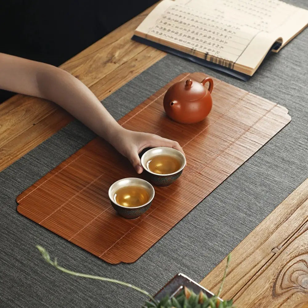 A hand-woven bamboo tea mat with heat insulation features a terracotta teapot and two small cups on a wooden table.