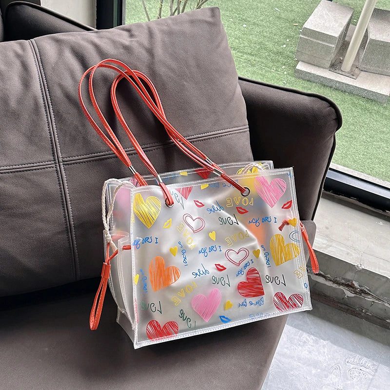 A clear transparent tote bag set with a mesh inner pouch and red drawstrings resting on a dark sofa.