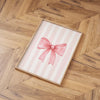 A framed canvas print featuring pink striped background and a pink bow on a wooden floor.