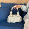 A hand is holding a white soft PU leather shoulder bag with silver rivet accents on a blue sofa.