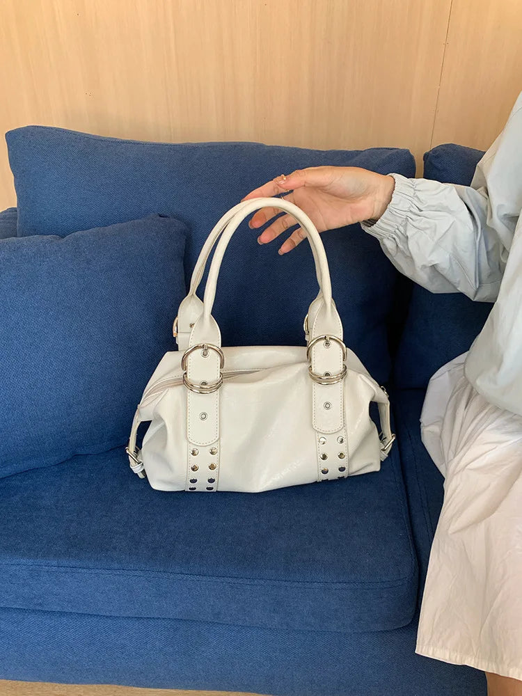 A hand is holding a white soft PU leather shoulder bag with silver rivet accents on a blue sofa.