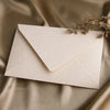 A cream colored linen texture envelope is placed on a silky beige fabric with a delicate sprig of dried foliage.