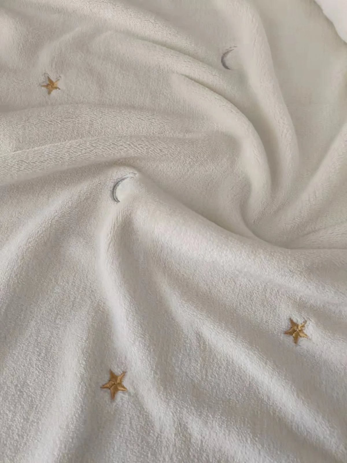 A close-up of a white coral fleece baby blanket with subtle gold star and crescent moon embroidery.