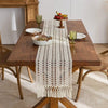A wooden dining table set for a meal with a beige tasseled table runner, plates, and glassware.