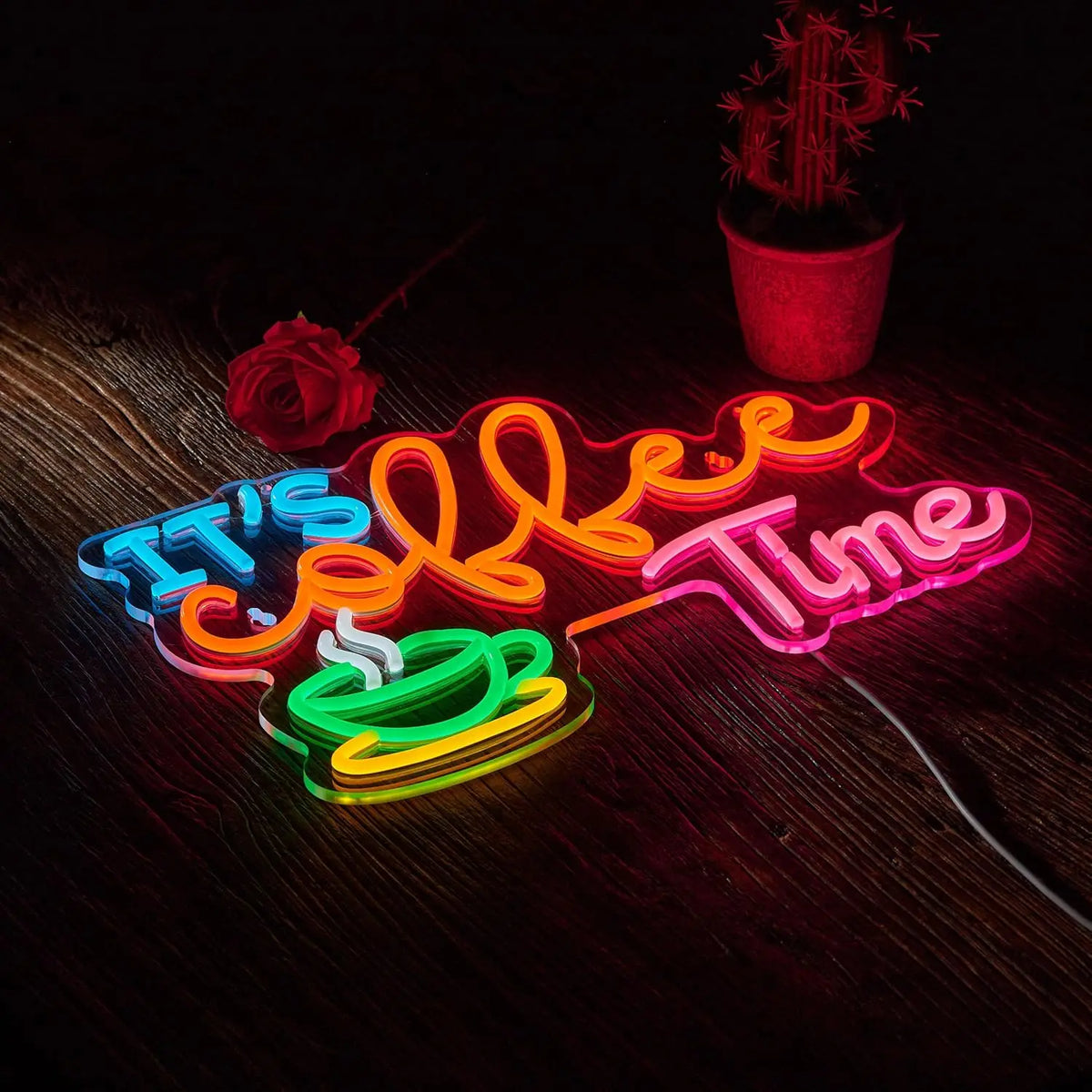 A neon sign shaped like 'It's Coffee Time' glows in various colors on a wooden surface next to a potted cactus.