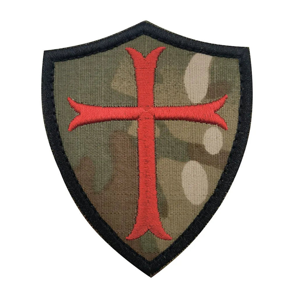 A dark olive green shield-shaped patch embroidered with a red cross, featuring hook and loop backing.