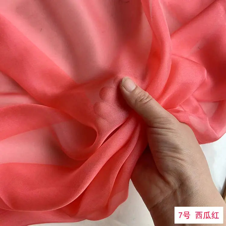 A hand is folding a piece of bright coral pink silk chiffon fabric to show its drape and texture.