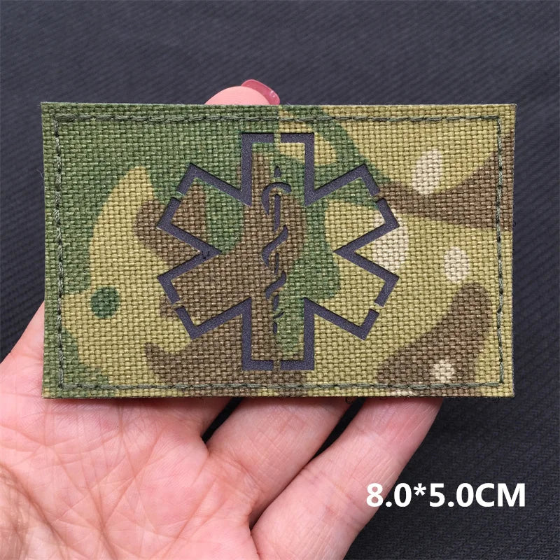 A rectangular olive green tactical patch featuring a dark gray medical cross design on a dark fabric background.