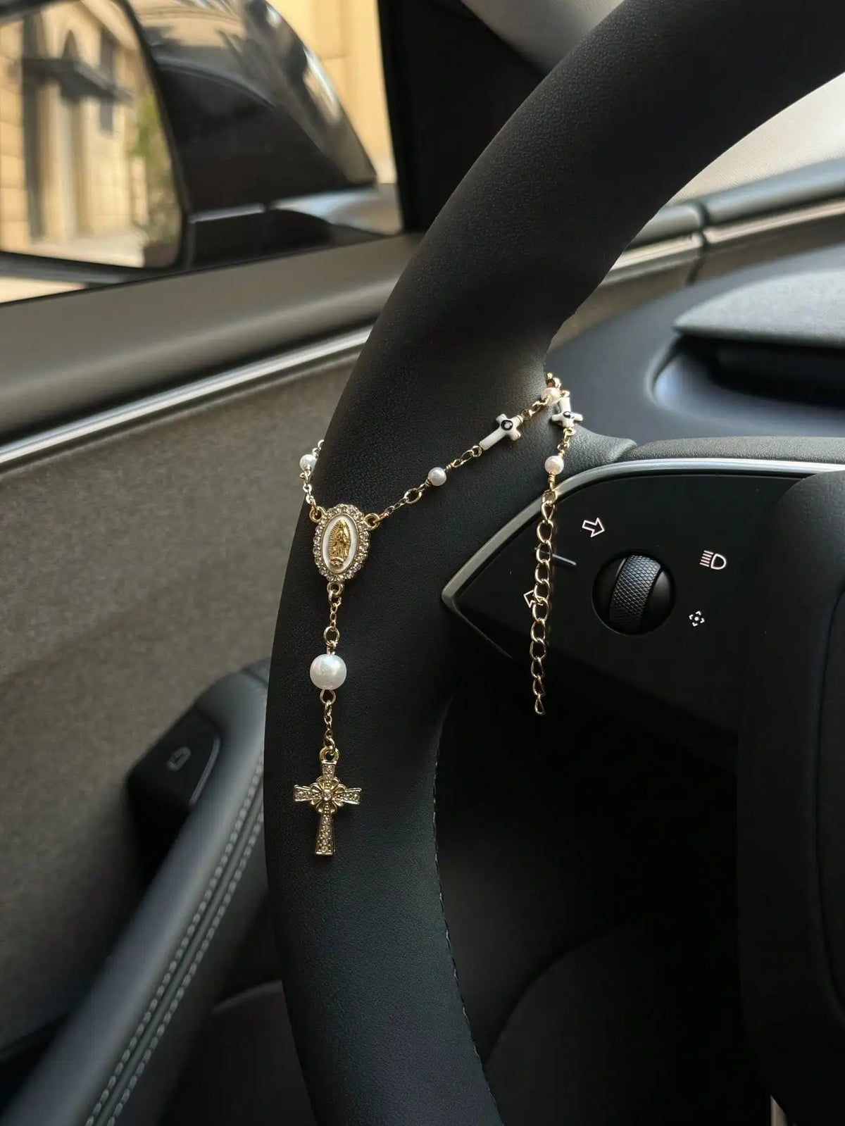 A gold rosary necklace with a pearl pendant is displayed on the steering wheel of a car.